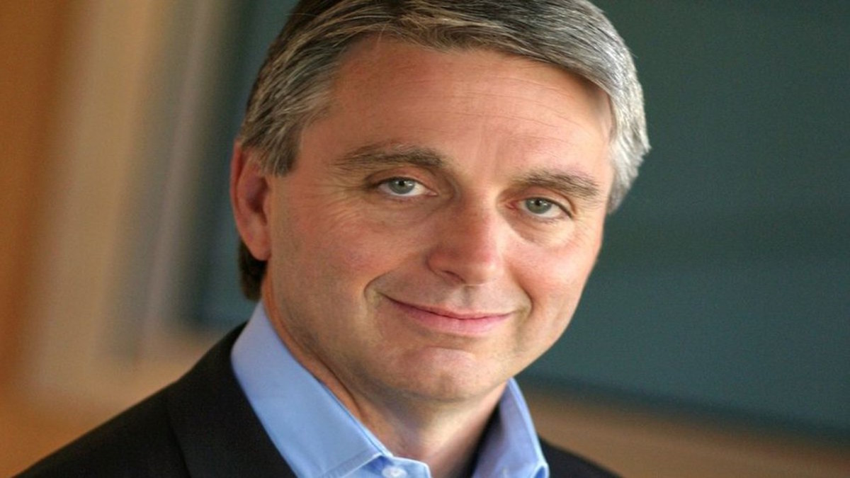 John Riccitiello, a white man with short silver hair and heavy eyes, is depicted wearing a jacket and collared shirt.